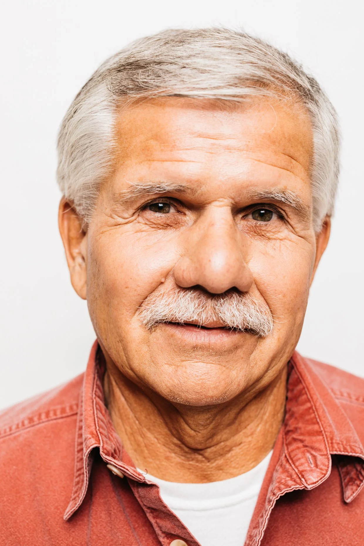 Close-up of an elderly man with gray hair, a mustache, wearing a salmon-colored button-up shirt, smiling softly against a plain white background.