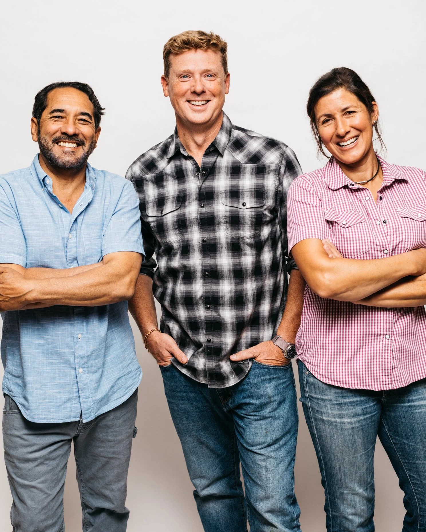 Three people standing together against a white background, smiling. The person on the left is a man with dark hair and a beard, wearing a light blue shirt. The person in the middle is a tall man with red hair, wearing a black and white plaid shirt. T