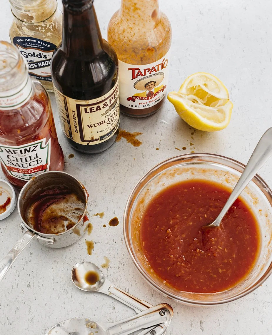 A bowl of homemade salsa with a spoon, surrounded by bottles of hot sauce, ketchup, and barbecue sauce, along with a halved lemon and measuring spoons on a light-colored surface.
