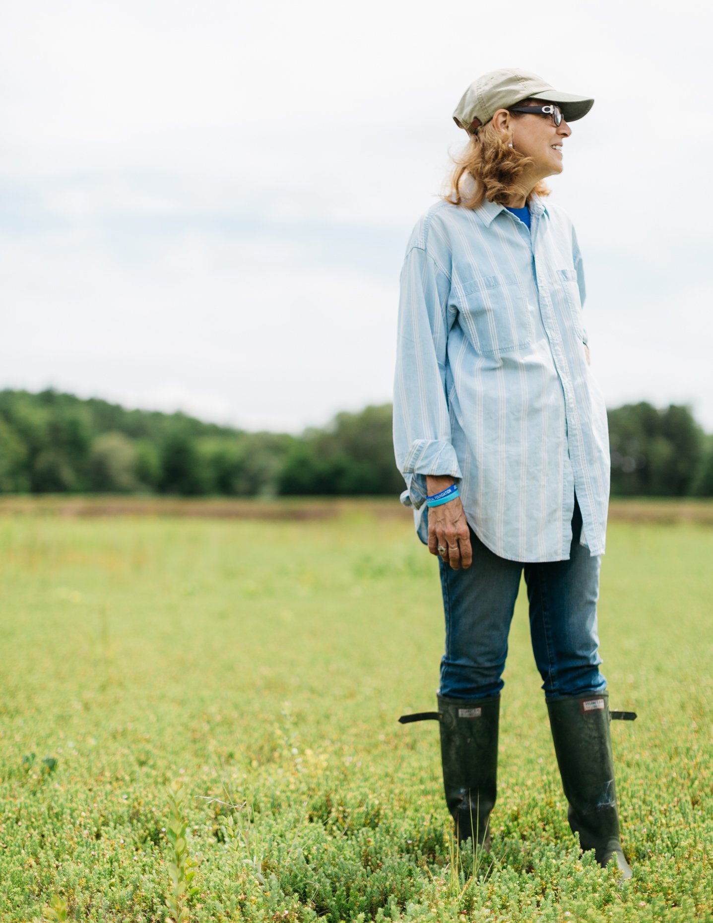 A woman stands in a field wearing a beige cap, sunglasses, a light blue button-up shirt, jeans, and black rubber boots, smiling while looking to her right.
