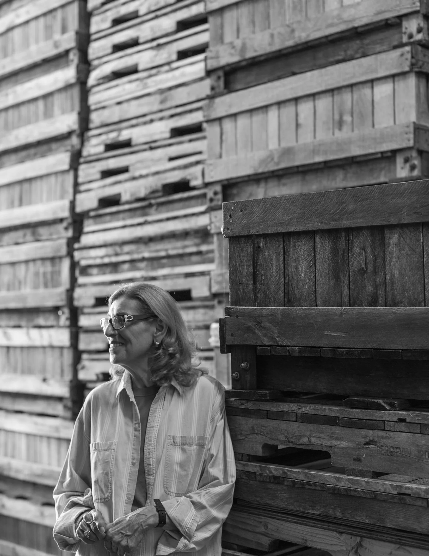 A woman with glasses and blonde hair smiles as she stands among stacked wooden crates in a warehouse.