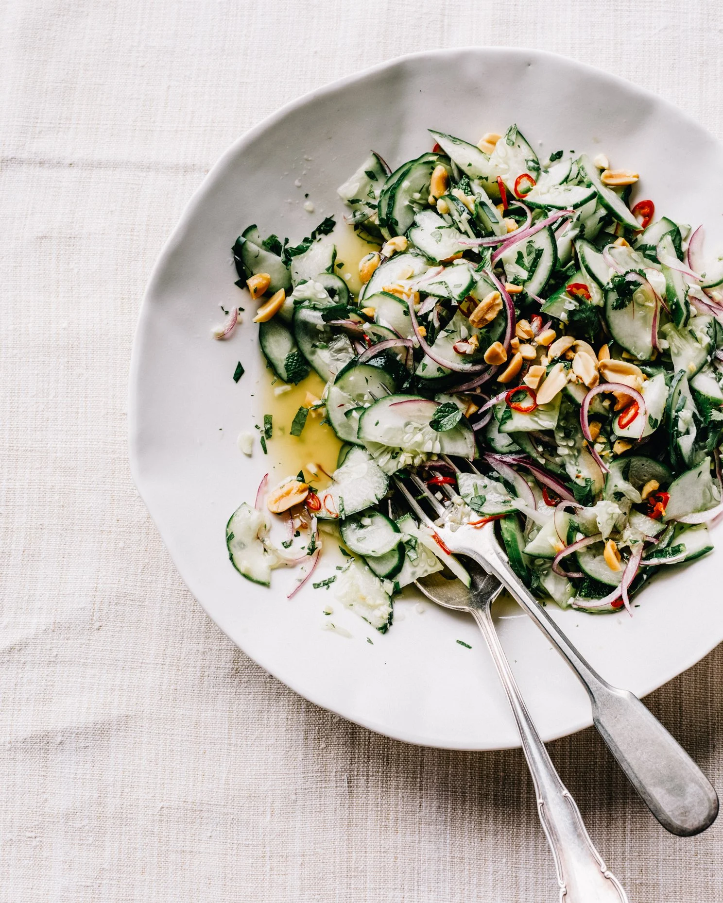 Cucumber salad with red onions, chopped herbs, peanuts, and sliced red chili on a white plate with a fork, placed on a beige tablecloth.