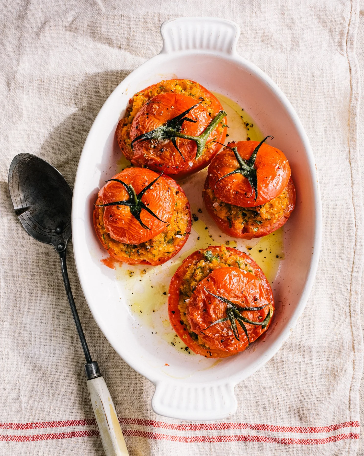 Baked stuffed tomatoes in a white oval dish with olive oil and herbs, on a beige napkin with a vintage spoon nearby.