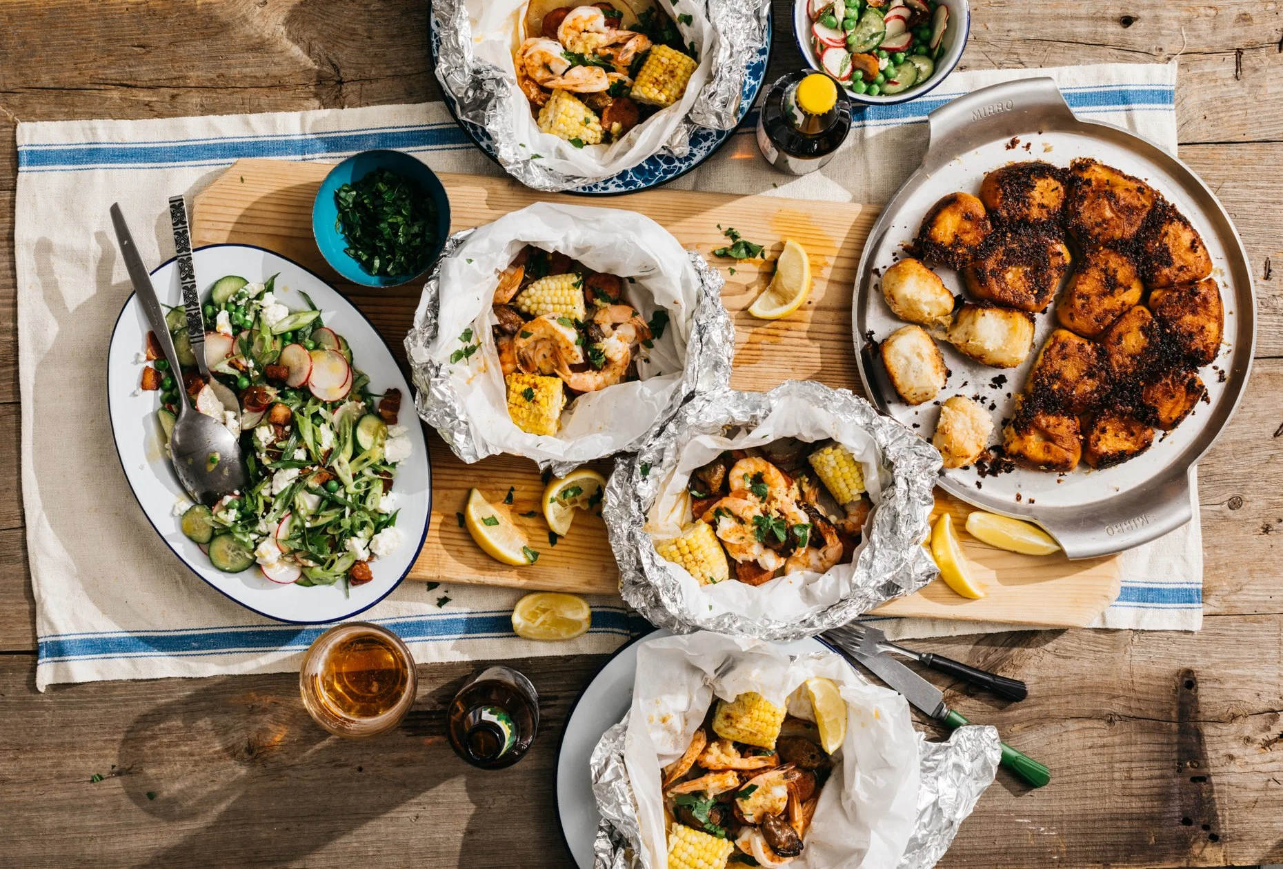 A rustic table setting with a variety of seafood dishes, including foil-wrapped lobster tails, grilled shrimp with corn on the cob, a garden salad, and a baked fish fillet, accompanied by lemon wedges, dipping sauces, and beverages.