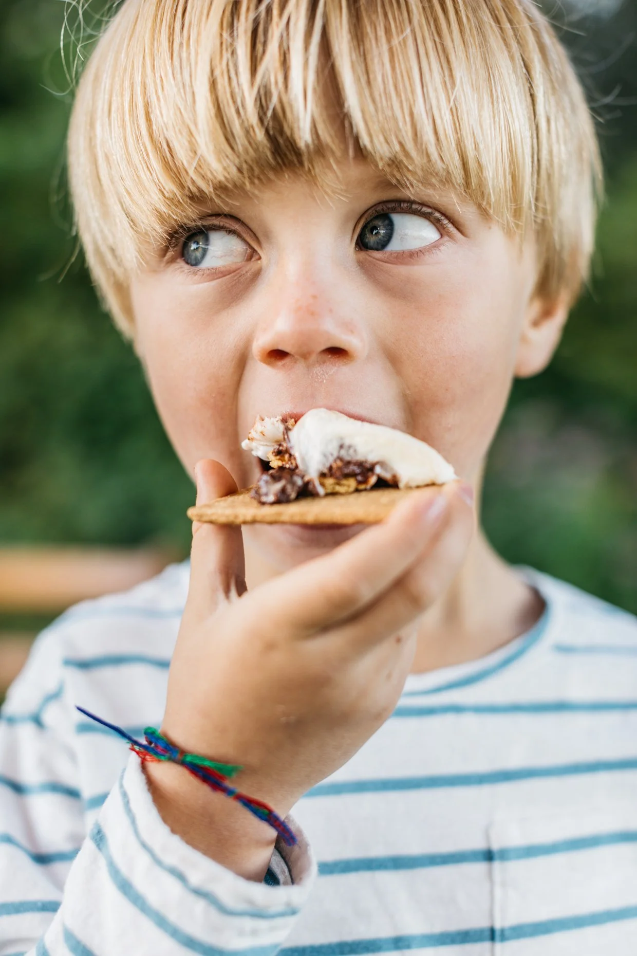 A young boy with blond hair and blue eyes eating a s'more outdoors.
