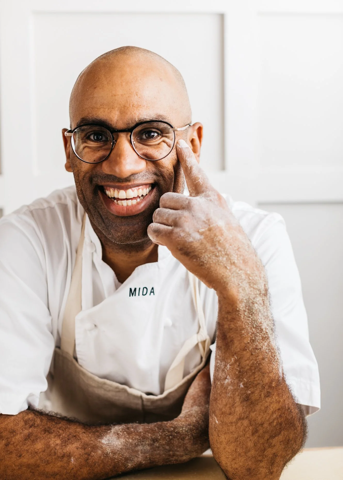A smiling man with glasses in a white chef's coat and apron, touching his face with a powdered hand, sitting at a table in a bright kitchen.