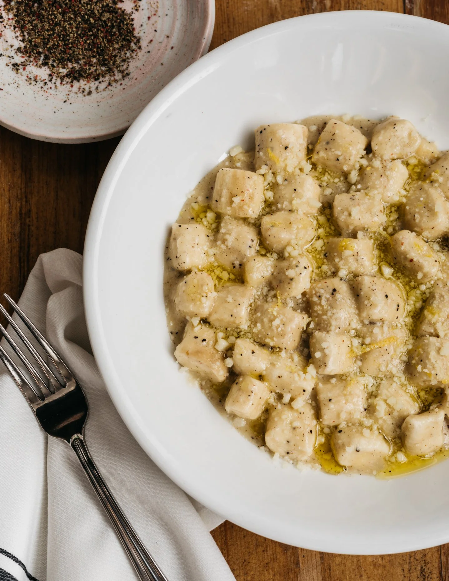 A white bowl filled with gnocchi in a creamy sauce, garnished with minced garlic and black pepper, placed on a wooden table near a fork, napkin, and a small bowl of ground black pepper.