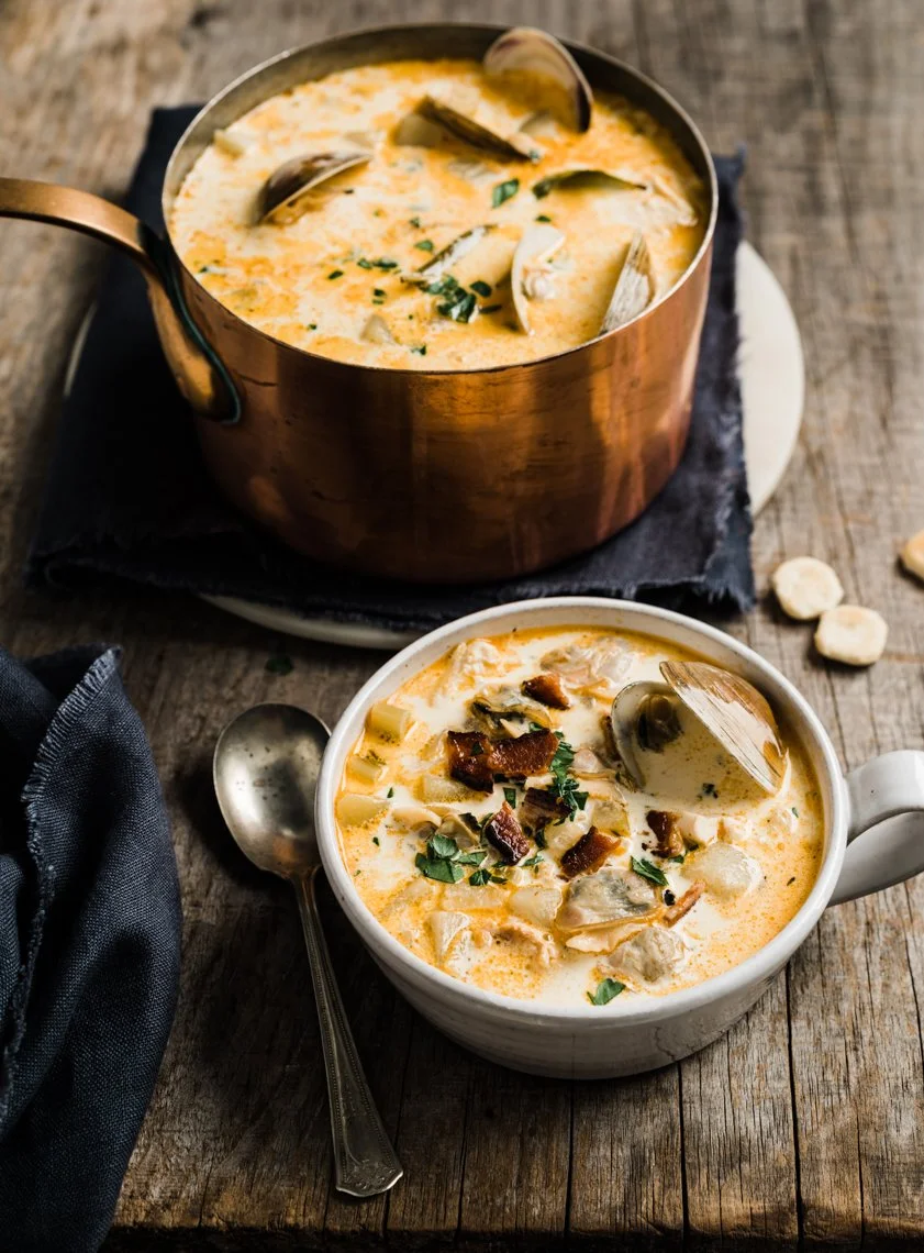 Two bowls of creamy seafood chowder with clams, bacon bits, and chopped herbs on a rustic wooden table, one in a copper pot and the other in a white mug.