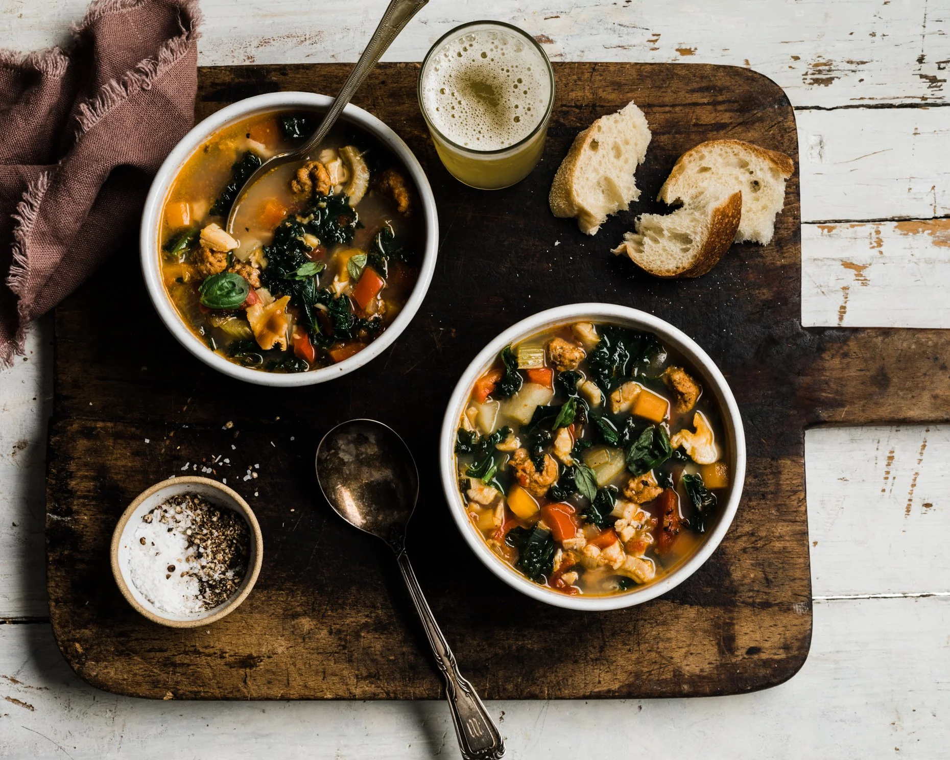 Two bowls of vegetable soup with carrots, kale, and squash, a glass of green juice, two pieces of sliced bread, a small bowl of salt and pepper, on a wooden cutting board.