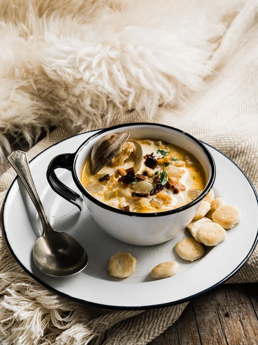A bowl of clam chowder with a clam shell on top, surrounded by oyster crackers on a white plate with a silver spoon, on a rustic wooden table with a cozy blanket in the background.