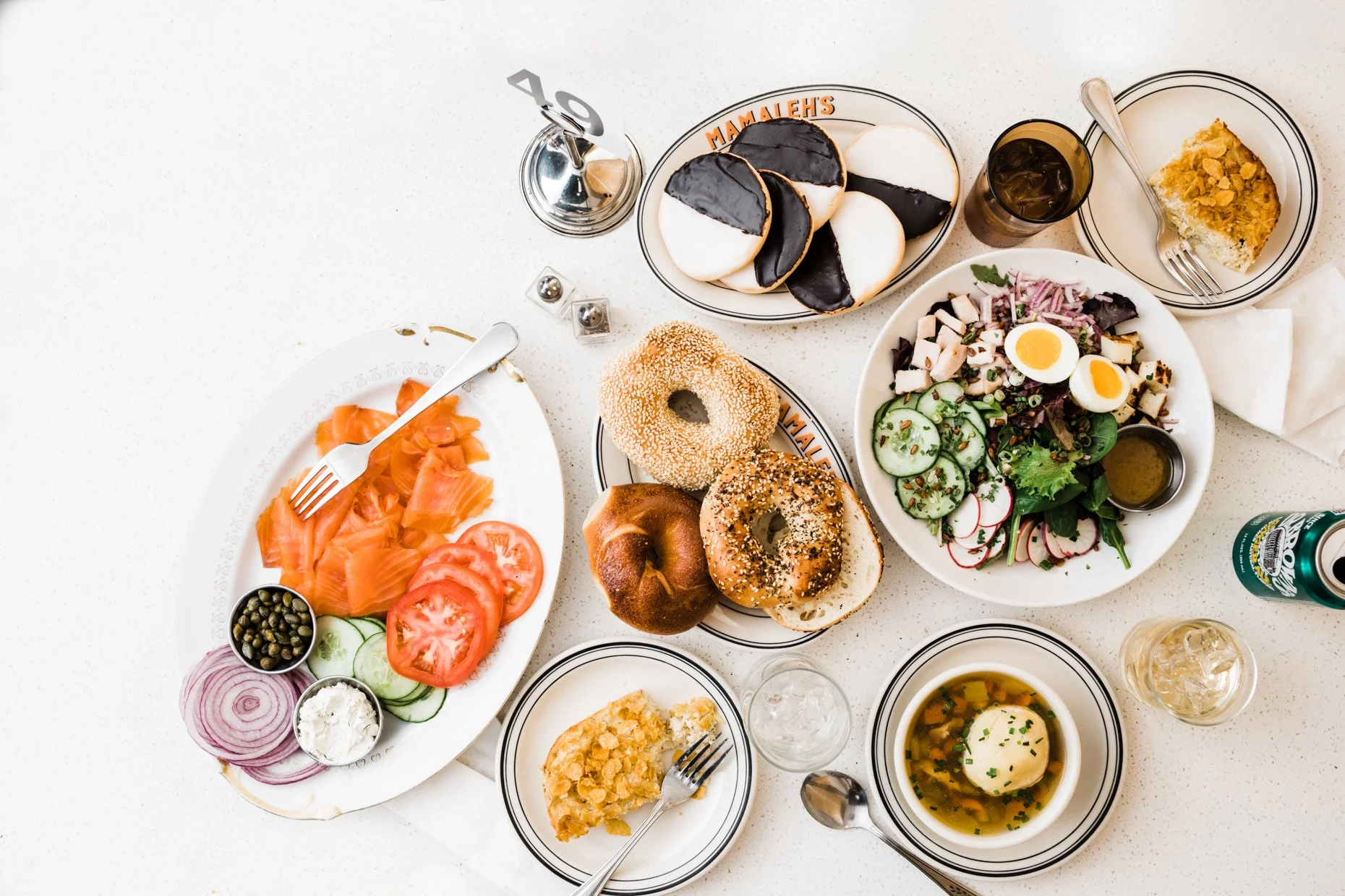 A table set with various dishes including smoked salmon with sliced cucumbers, tomatoes, red onion, capers, and cream cheese; mixed salad with boiled eggs, cucumbers, radishes, and leafy greens with dressing; a bowl of soup with a hard-boiled egg; tw