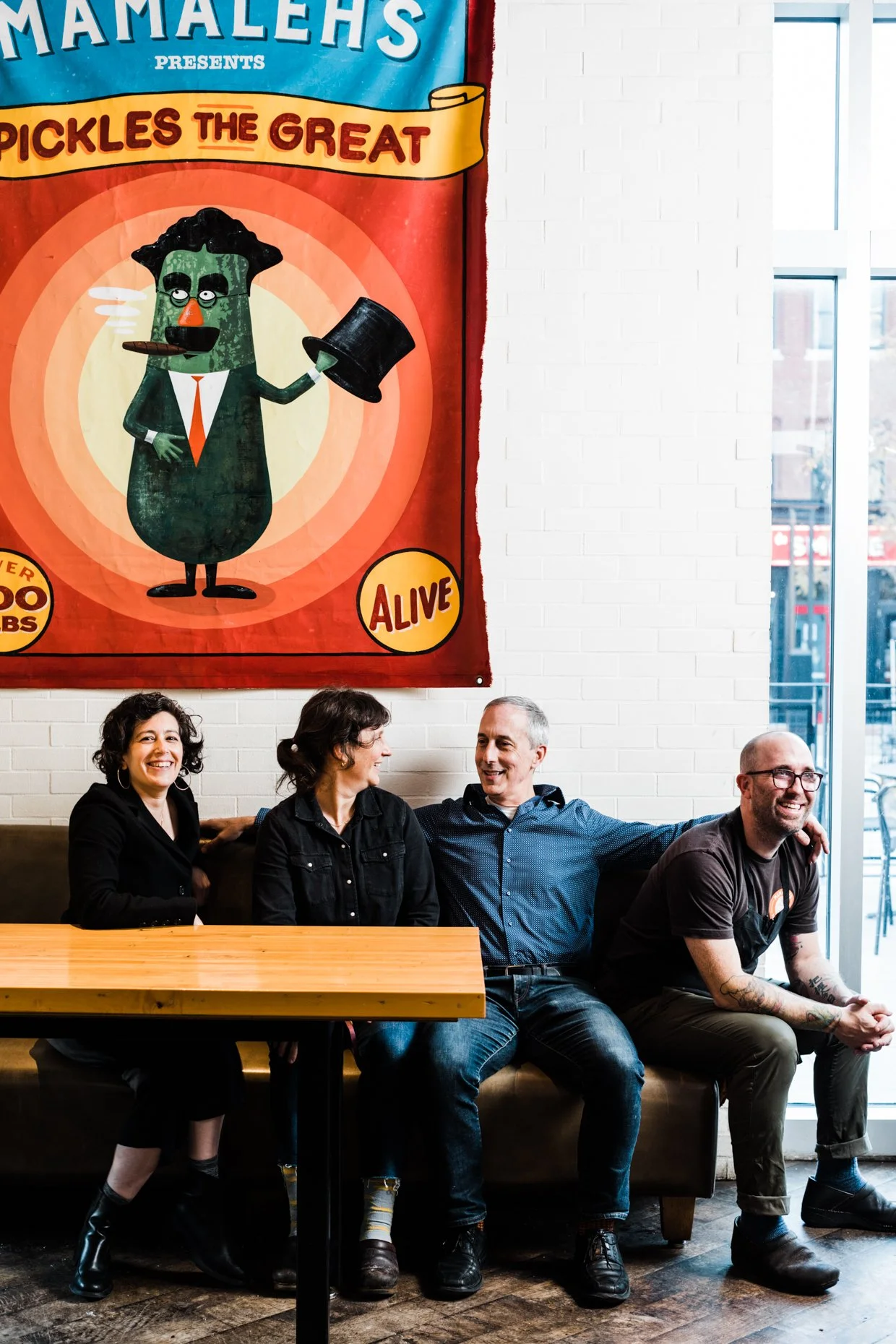 Four people sitting on a couch chatting and laughing in a cafe with a colorful mural of a pickle dressed in a suit and holding a top hat on the wall behind them.