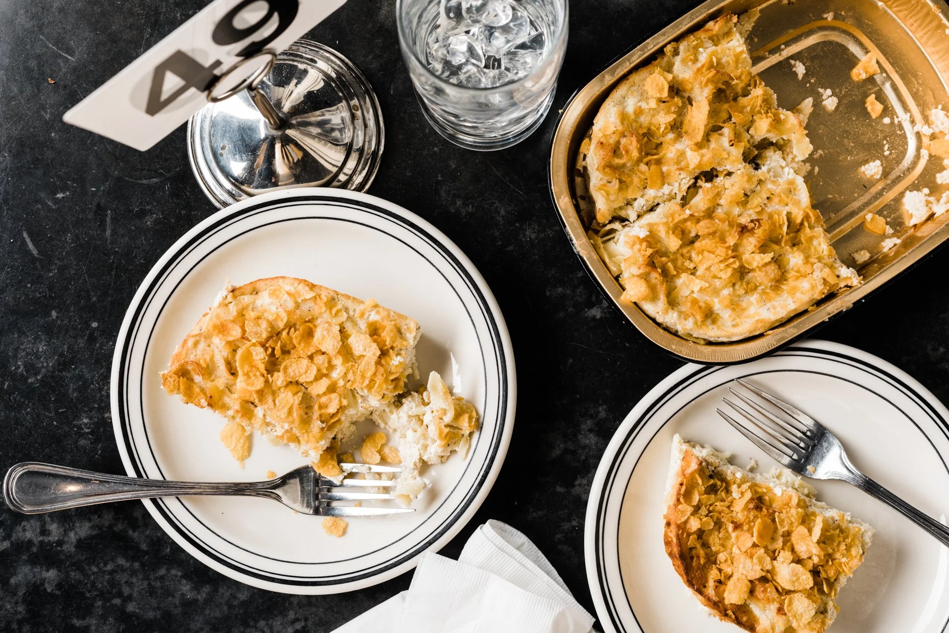 Slices of apple crumble pie on white plates with forks, a metal container with more pie, a glass of ice water, and a table number sign on a black marble table.