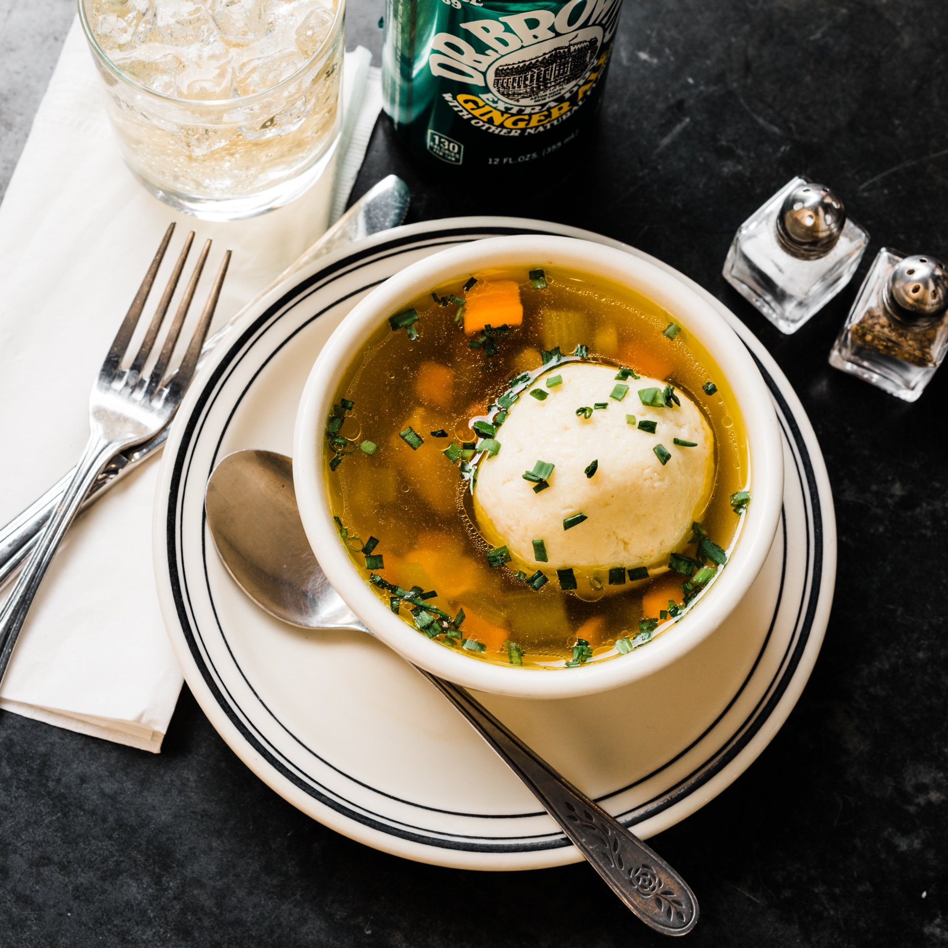 A bowl of hot chicken noodle soup topped with a scoop of mashed potatoes, garnished with chopped chives, on a white plate with a spoon. Accompanied by a glass of ice water, a can of Dr. Bronner's ginger ale, and salt and pepper shakers on a black tab