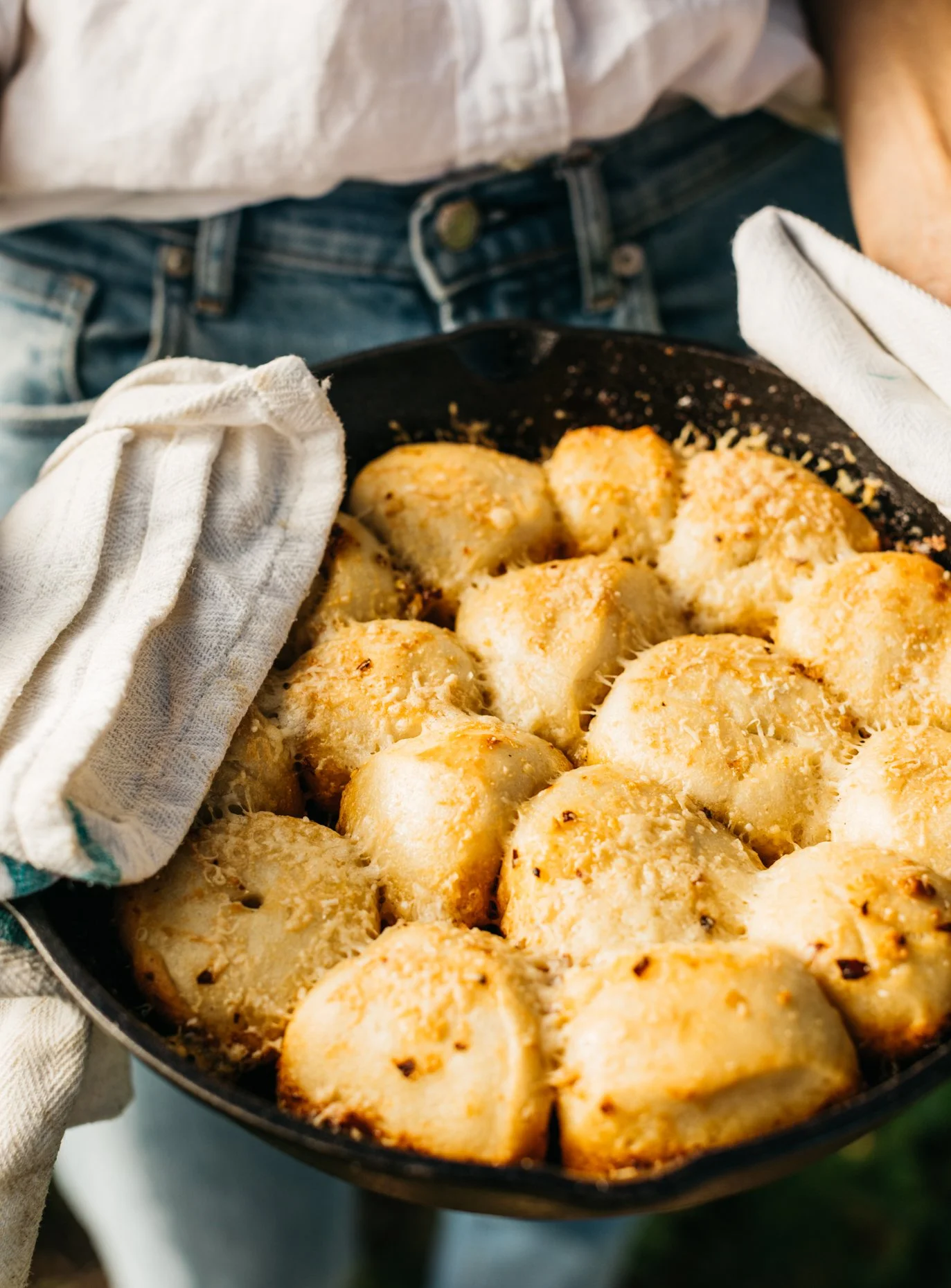 Person holding a cast iron skillet filled with golden-brown baked bread rolls, using oven mitts.