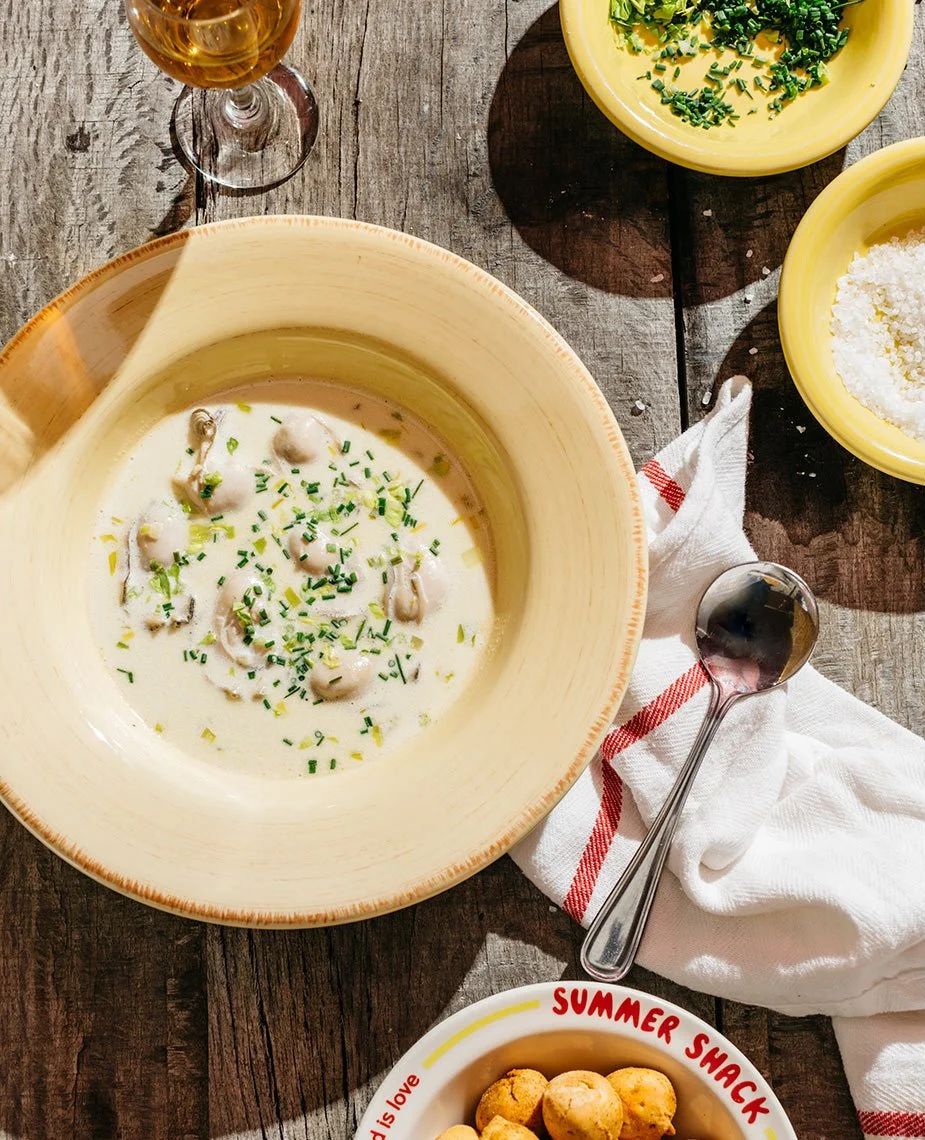 A rustic wooden table with a bowl of creamy mushroom soup garnished with chopped chives, a glass of white wine, two yellow bowls with chopped herbs and grated cheese, a silver soup spoon on a white cloth, a small bowl of summer snacks, and a napkin w