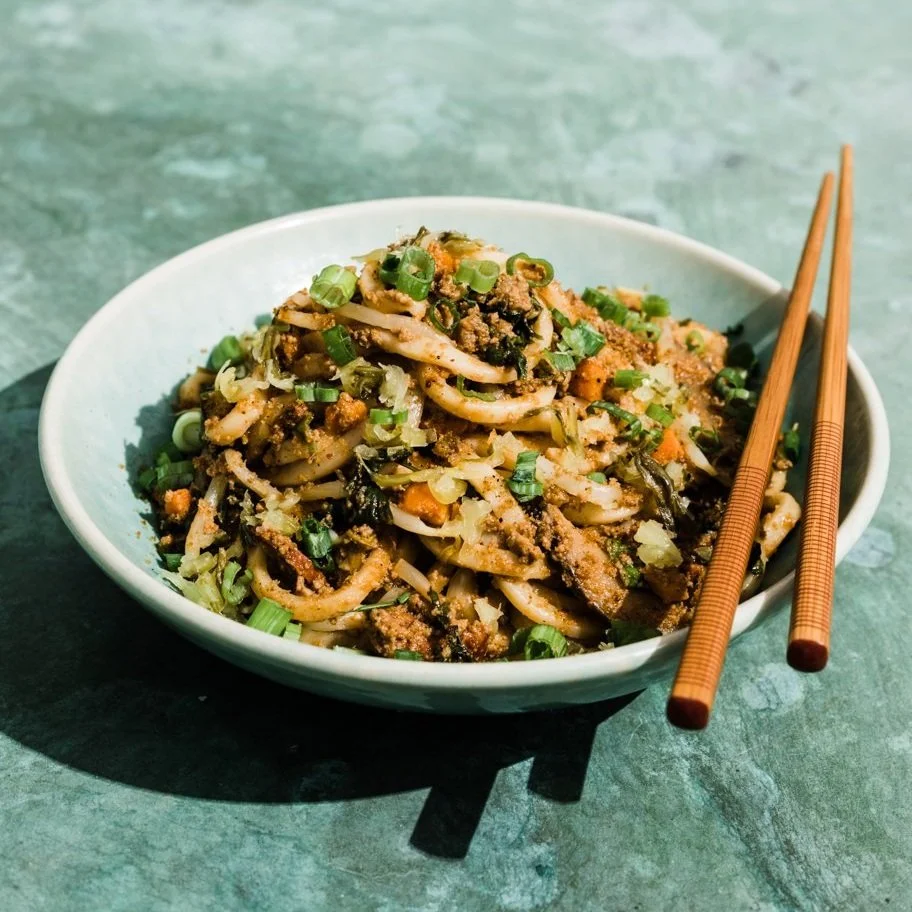 Bowl of Asian-style stir-fried noodles with ground meat, chopped green onions, and vegetables, accompanied by chopsticks.