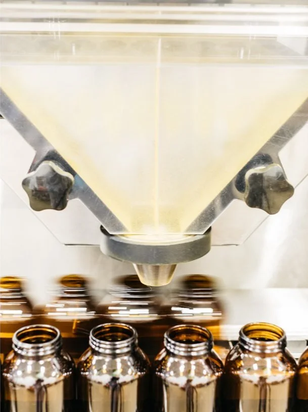 Close-up of a fill-funnel dispensing vitamins into amber glass bottles.