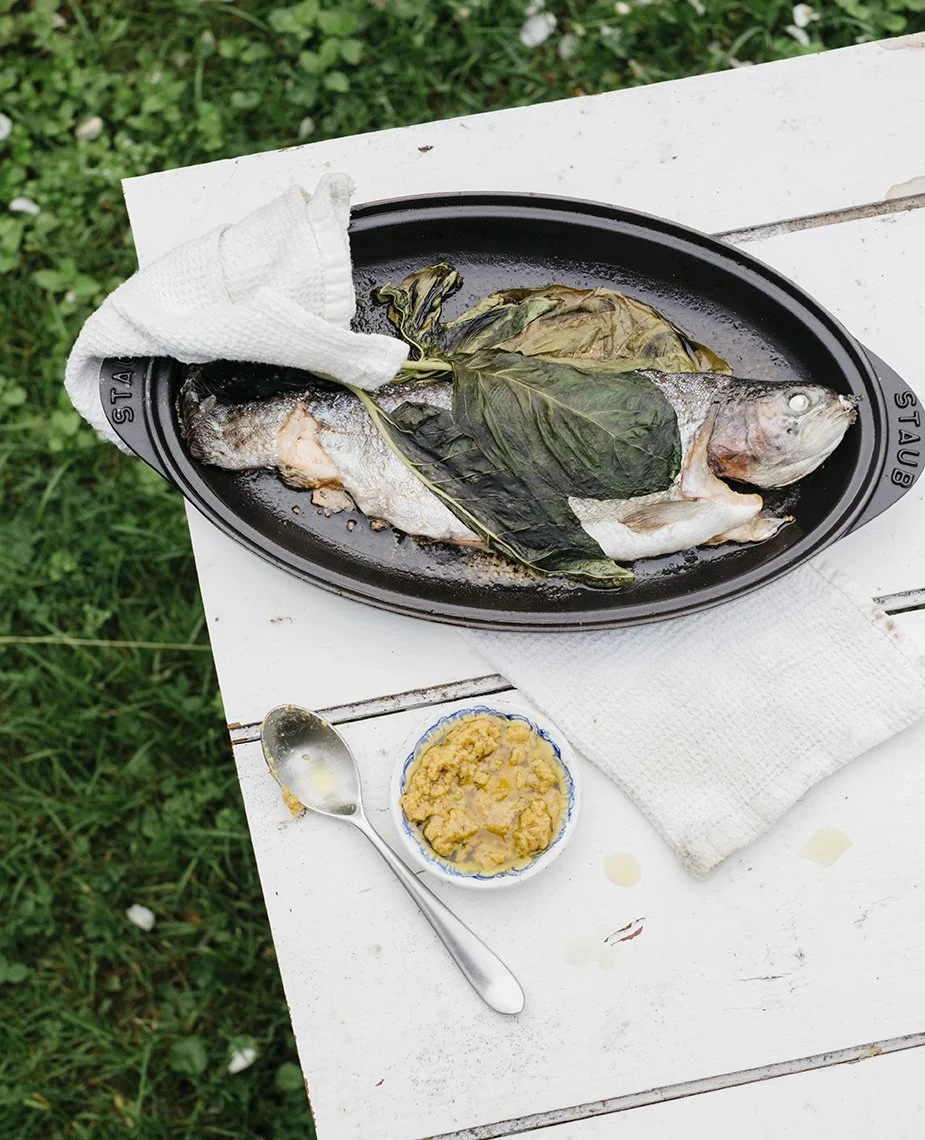 Whole fish cooked on a black oval baking dish, garnished with wilted leaves, on a white table outdoors, accompanied by a bowl of yellowish sauce and a silver spoon.