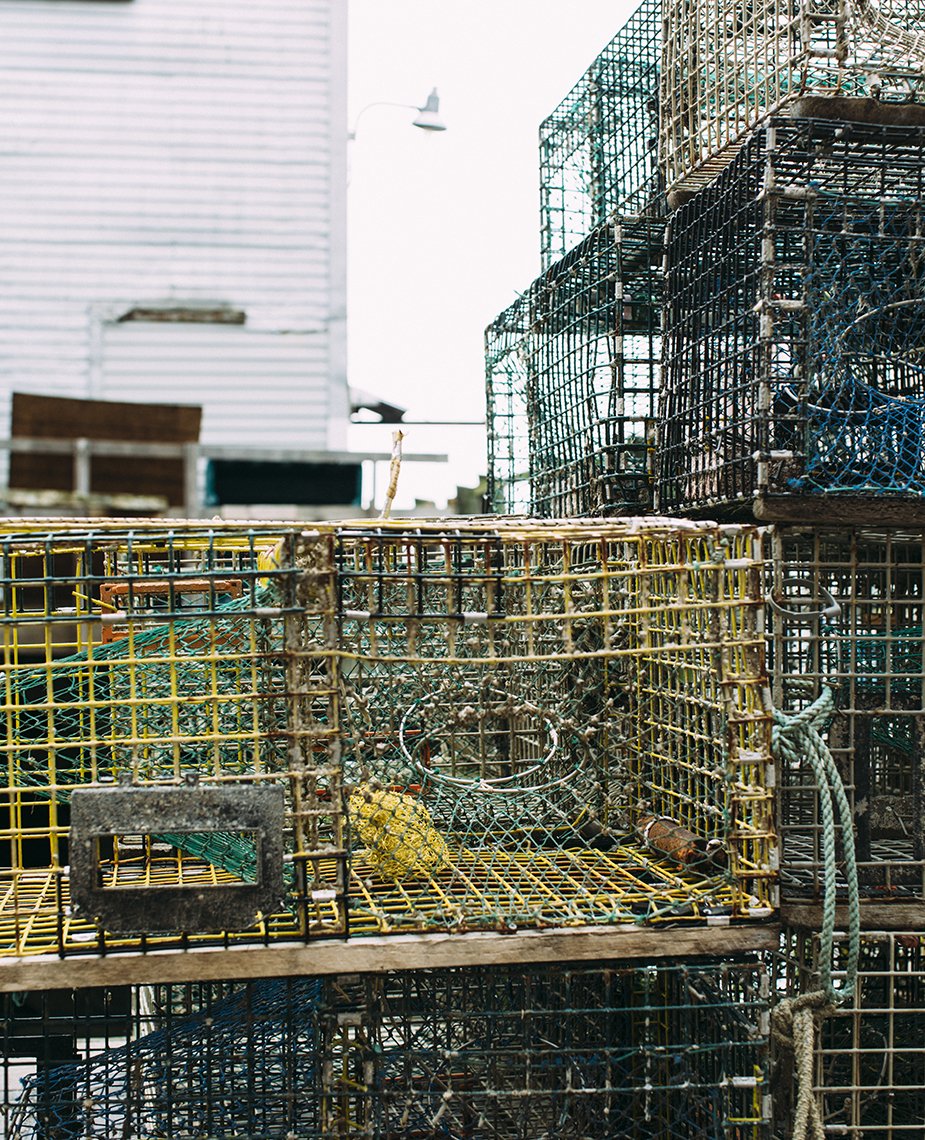Stacked crab traps made of metal wire in various colors including yellow, green, blue, and brown, with some ropes and a yellow float, outdoors against a cloudy sky and a building.