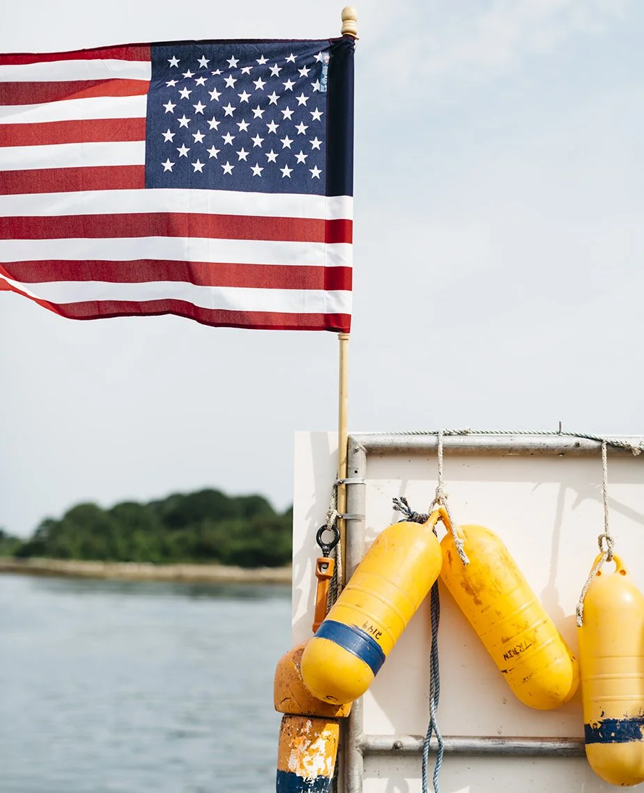 American flag on a pole flying above a boat with yellow buoys hanging on the side, water and greenery in the background.