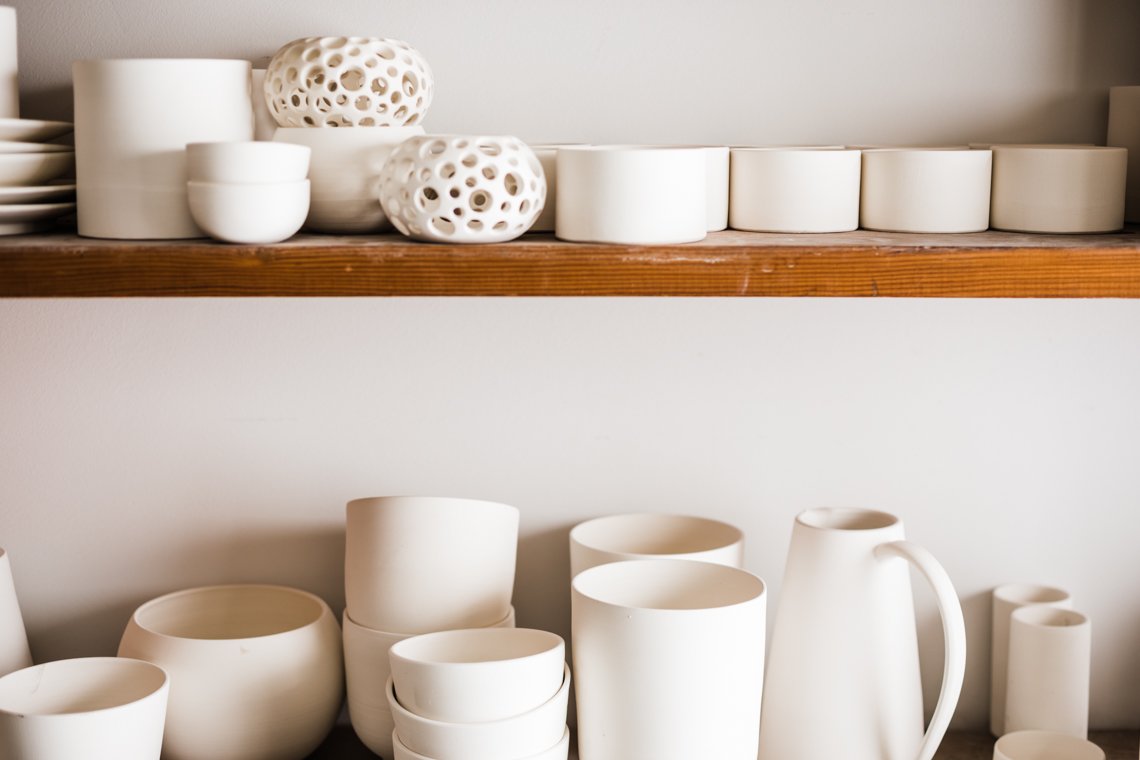 White ceramic bowls, mugs, and decorative vases placed on a wooden shelf and lower surface.
