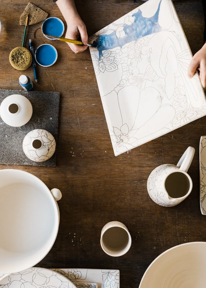Person painting a floral design on a ceramic plate with blue glaze, surrounded by white ceramic pottery, brushes, sponges, glue, and socks on a wooden table.