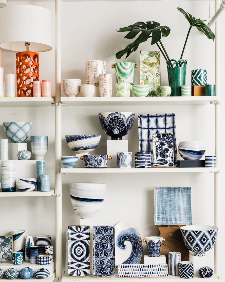 Display of various ceramic and glass bowls, cups, and vases with patterns in blue, white, and green on white shelves. There is a large green plant in a green vase on the top right.