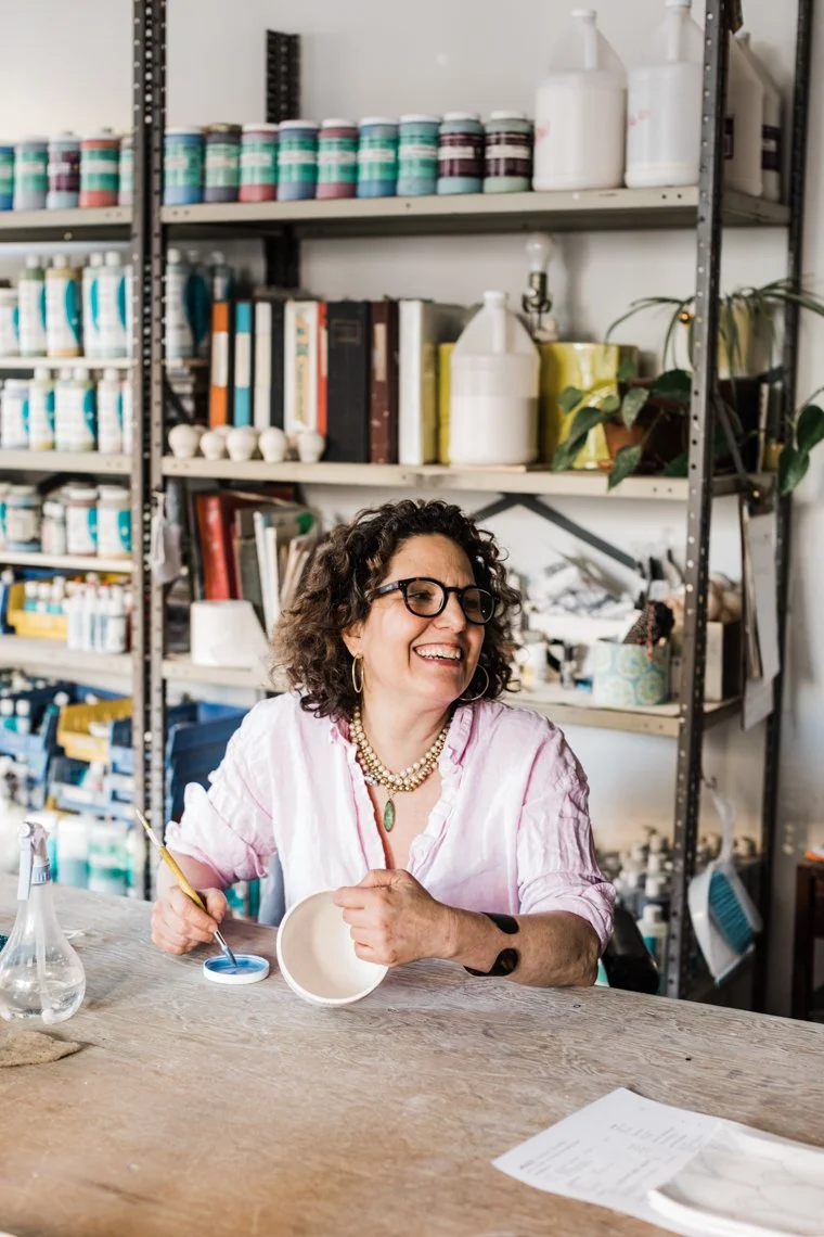A woman with curly hair and glasses sitting at a work table, painting a ceramic bowl with blue glaze, surrounded by shelves with ceramic supplies and books.