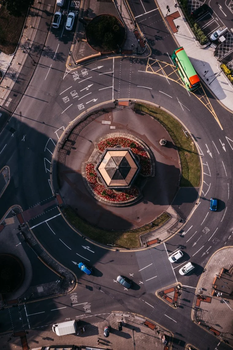 An aerial view of a circular roundabout with a small central park with flowers, surrounded by multiple lanes of traffic with cars and a green bus. The streets are marked with arrows and directional signs.
