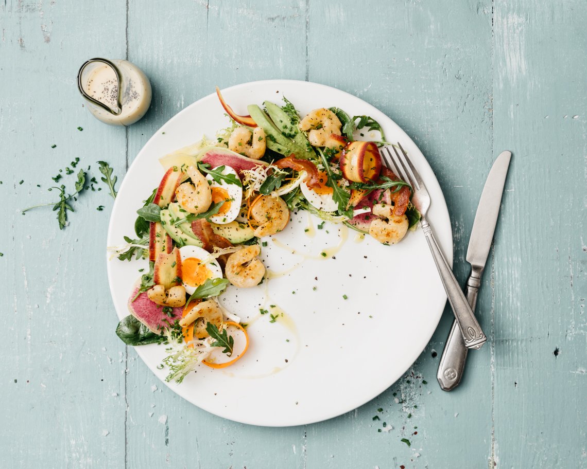 Composed salad, including plant-based shrimp, boiled eggs, cucumber, and mixed greens, on a light blue wooden table with a fork and knife beside it and a small container of dressing nearby.
