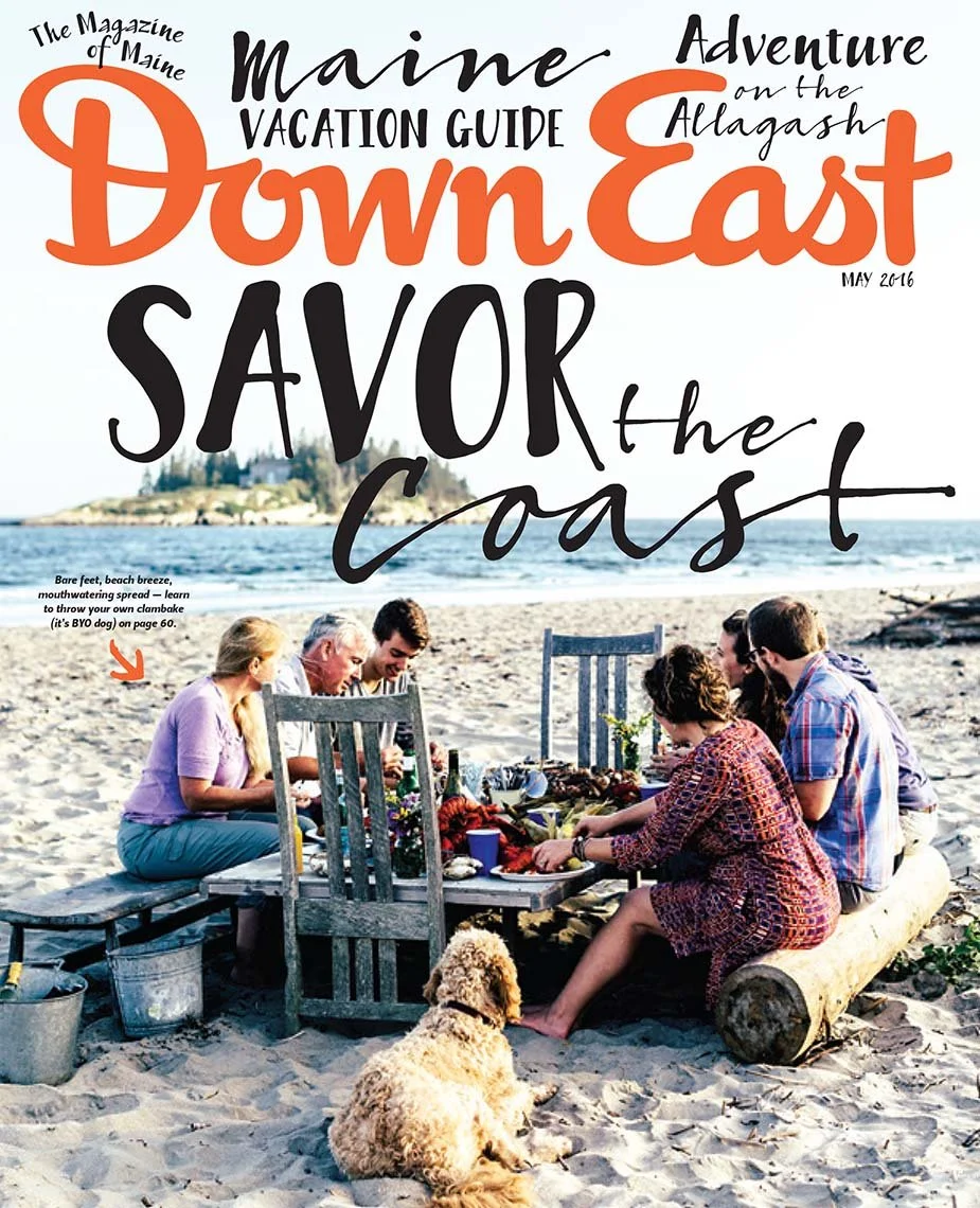 A group of people having a beach picnic on the sand near the ocean with a dog sitting nearby, surrounded by chairs, a table, and picnic items. The photo is overlaid with colorful text advertising a Maine vacation guide.