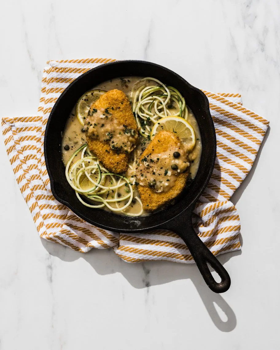 Fried fish fillets with lemon slices in a creamy sauce, garnished with herbs and black pepper, served with spiralized zucchini noodles in a black cast iron skillet on a white marble surface with a striped cloth underneath.