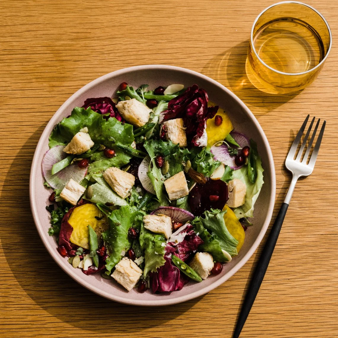 A bowl of mixed salad with leafy greens, radicchio, cucumber, pomegranate seeds, croutons, and a yellow dressing, on a wooden table with a glass of yellow beverage and a fork.