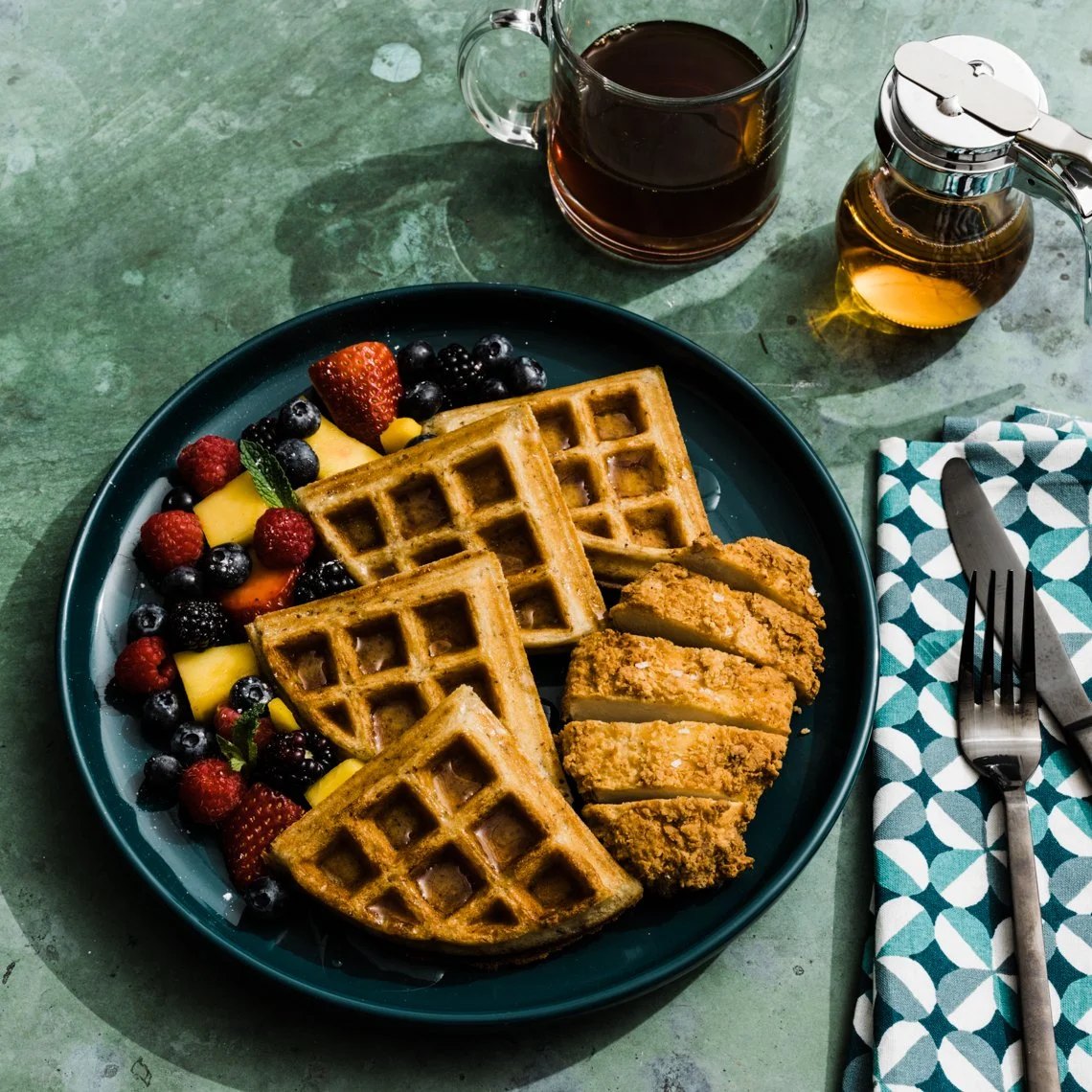 A plate with waffles, fried chicken strips, and mixed berries on a green table, with a glass of dark coffee, a honey jar, and a knife and fork on a blue patterned napkin.