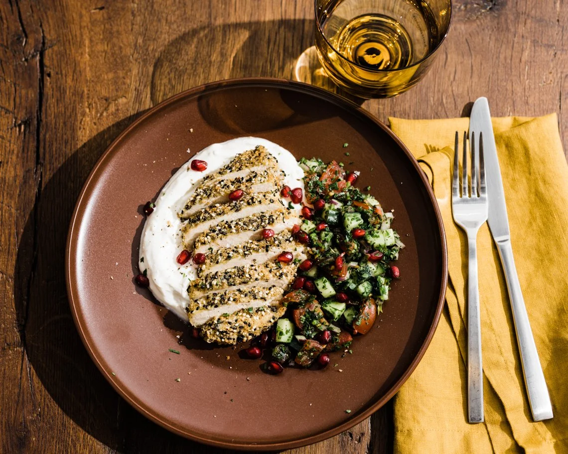 A plate of herb-crusted chicken breast on a bed of creamy sauce, served with cucumber and tomato salad garnished with pomegranate seeds, with a glass of white wine on a wooden table
