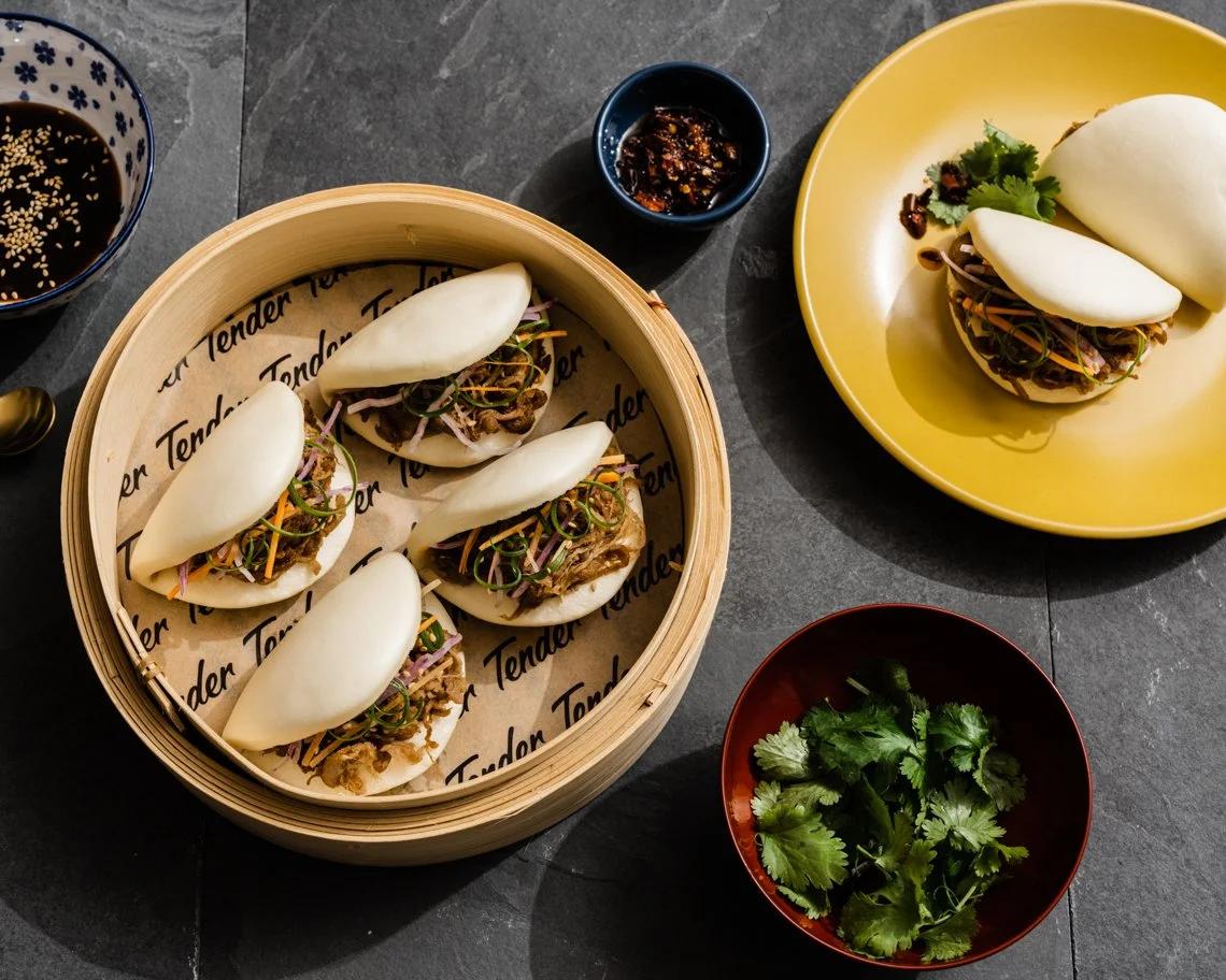 Six steamed bao buns filled with shredded beef and vegetables, served in a bamboo steamer, with dipping sauce, soy sauce, and fresh cilantro on the side.