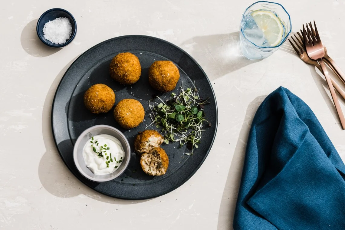 A black plate with five croquetas, a small bowl of creamy sauce with chopped chives, and a small bunch of microgreens. There is a glass of water with lemon, a small bowl of coarse salt, and a set of rose gold forks and knives on a white table with a 