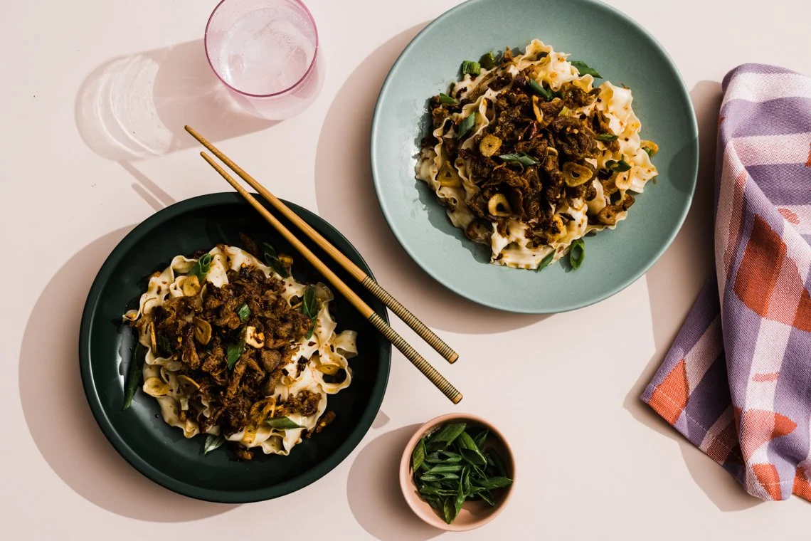 Two plates of cooked noodles with beef and scallions on a light-colored table, accompanied by a glass of water, a small bowl of chopped green onions, a pair of wooden chopsticks, and a folded cloth napkin.
