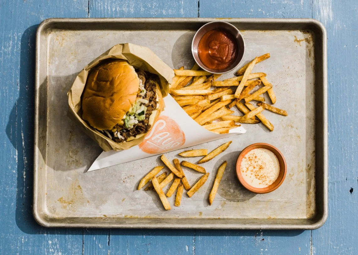 Leaning Tower of a cheeseburger with lettuce, on a piece of deli paper on a metal tray. Next to the burger are French fries, ketchup, and a side of creamy sauce on a terracotta-colored dish. The tray is on a blue wooden surface.