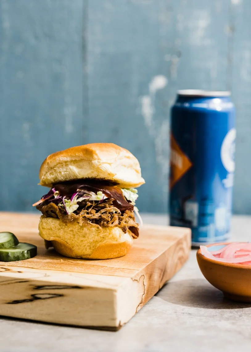 Pulled pork sandwich with coleslaw on a bun, sliced cucumbers, a can of soda, and a bowl of sliced pickles on a wooden cutting board.
