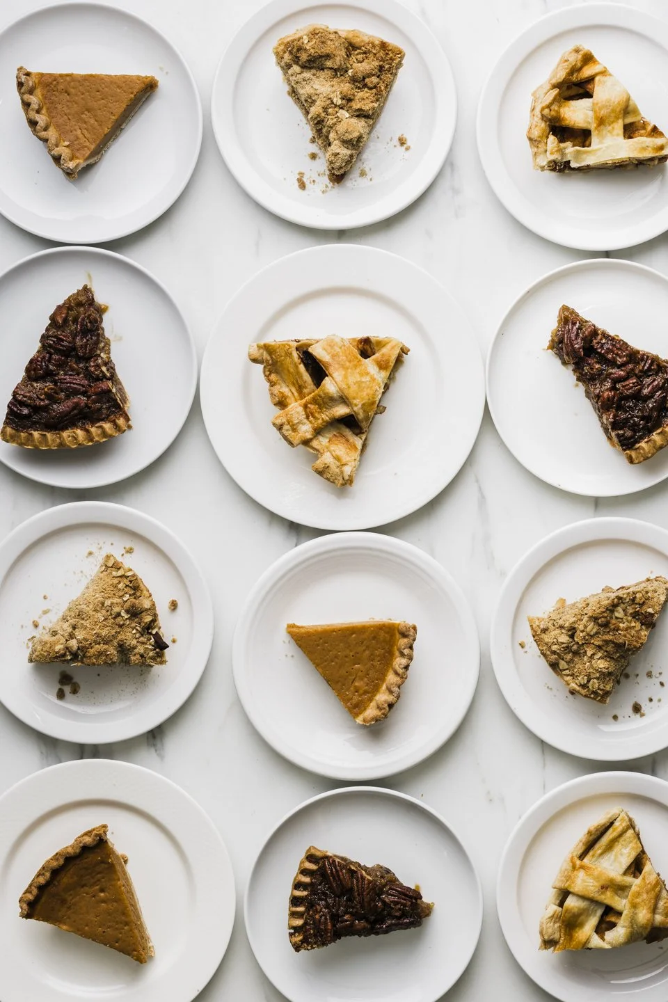 Assorted slices of pie arranged on white plates on a marble surface, including pumpkin, apple, cherry, pecan, and crumb-topped varieties.