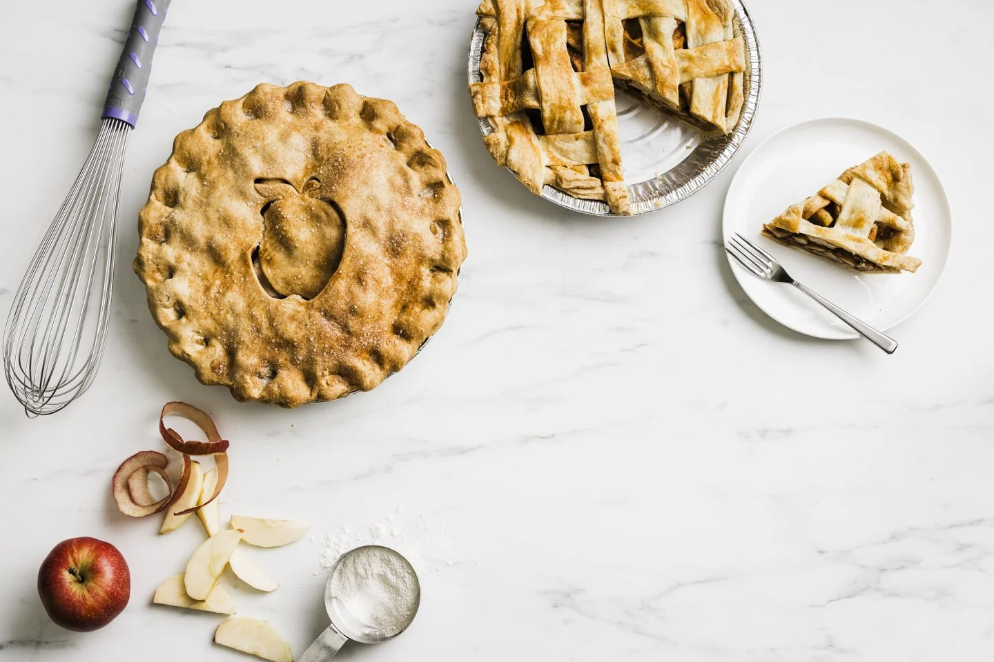 A freshly baked apple pie with a lattice crust next to a sliced pie on a white marble surface. There is a whole apple, apple peels, and a scoop of flour nearby, along with a whisk. A slice of pie is on a white plate with a fork, and additional slices