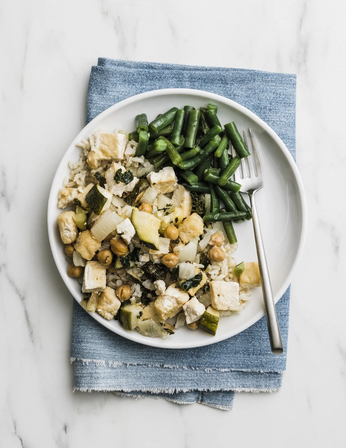 Plate of rice with chickpeas, tofu, zucchini, and greens, with green beans on the side, on a white marble surface with a blue napkin and fork.