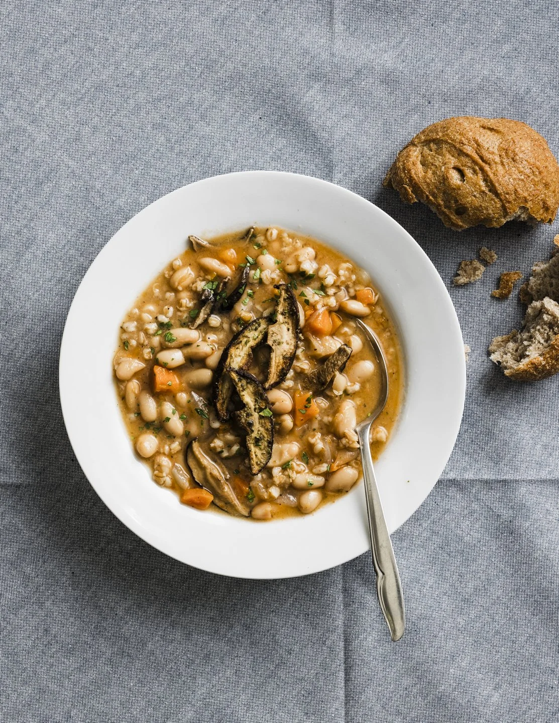 White bowl of vegetable and bean soup topped with fried eggplant slices, on a gray tablecloth with a piece of bread and a muffin nearby