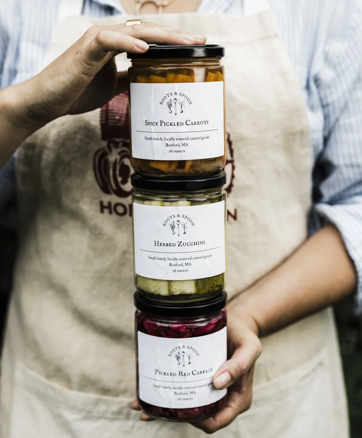 Person holding three stacked jars of canned vegetables labeled 'Spicy Pickled Carrots,' 'Herbed Zucchini,' and 'Pickled Red Cabbage' from Roots & Spoon in Boxford, MA.