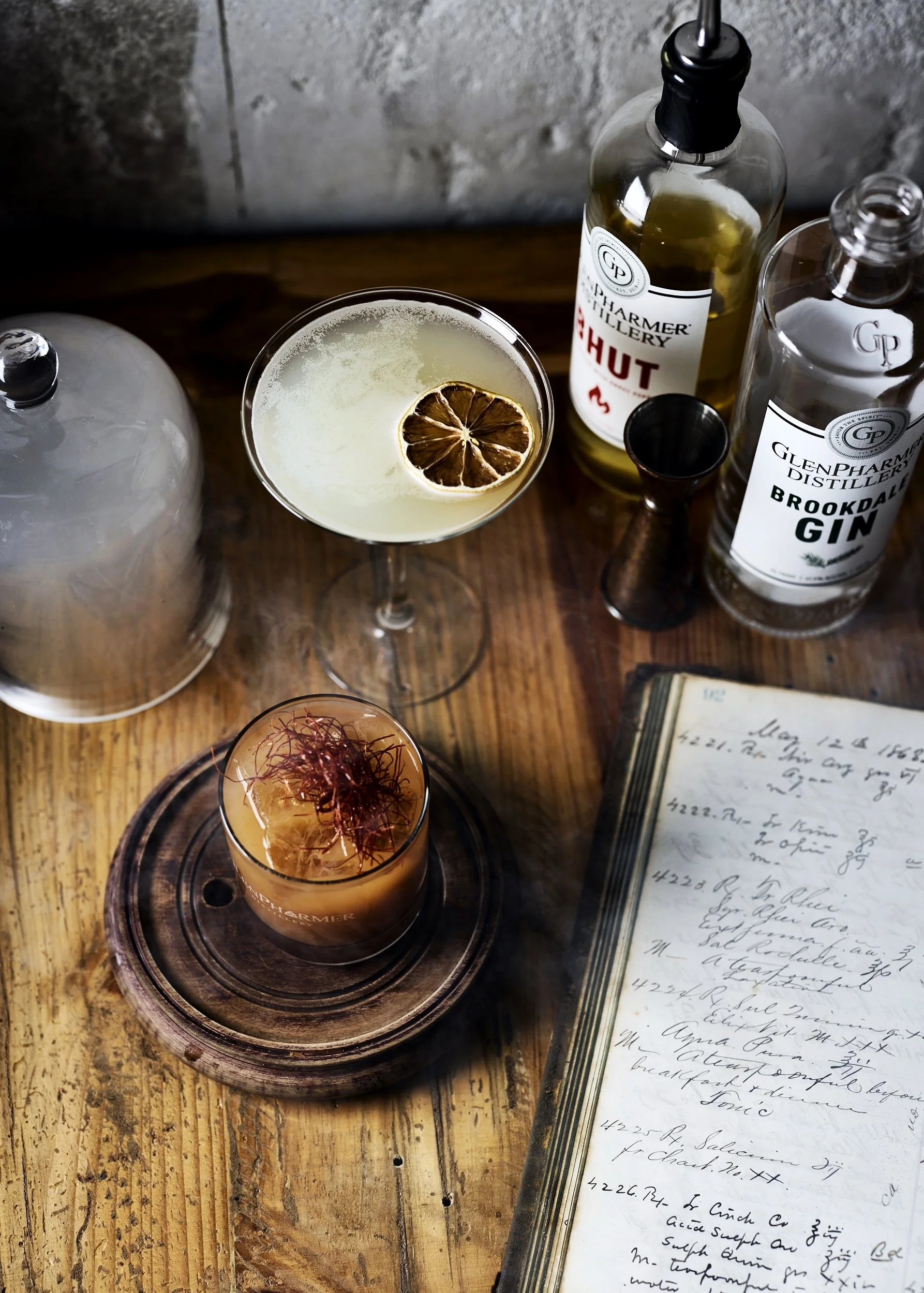 A wooden table with a cocktail garnished with a dried lemon slice, a glass with dark garnish, a flip top bottle labeled 'GlenPharmer Distillery Brookdale Gin', and a handwritten recipe journal.