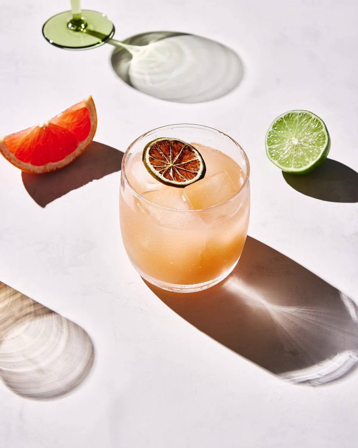 A grapefruit and a lime quarter on a white surface along with a pink cocktail in a glass with ice and a dried lime slice as garnish. Shadows cast by the glass and citrus pieces are visible.
