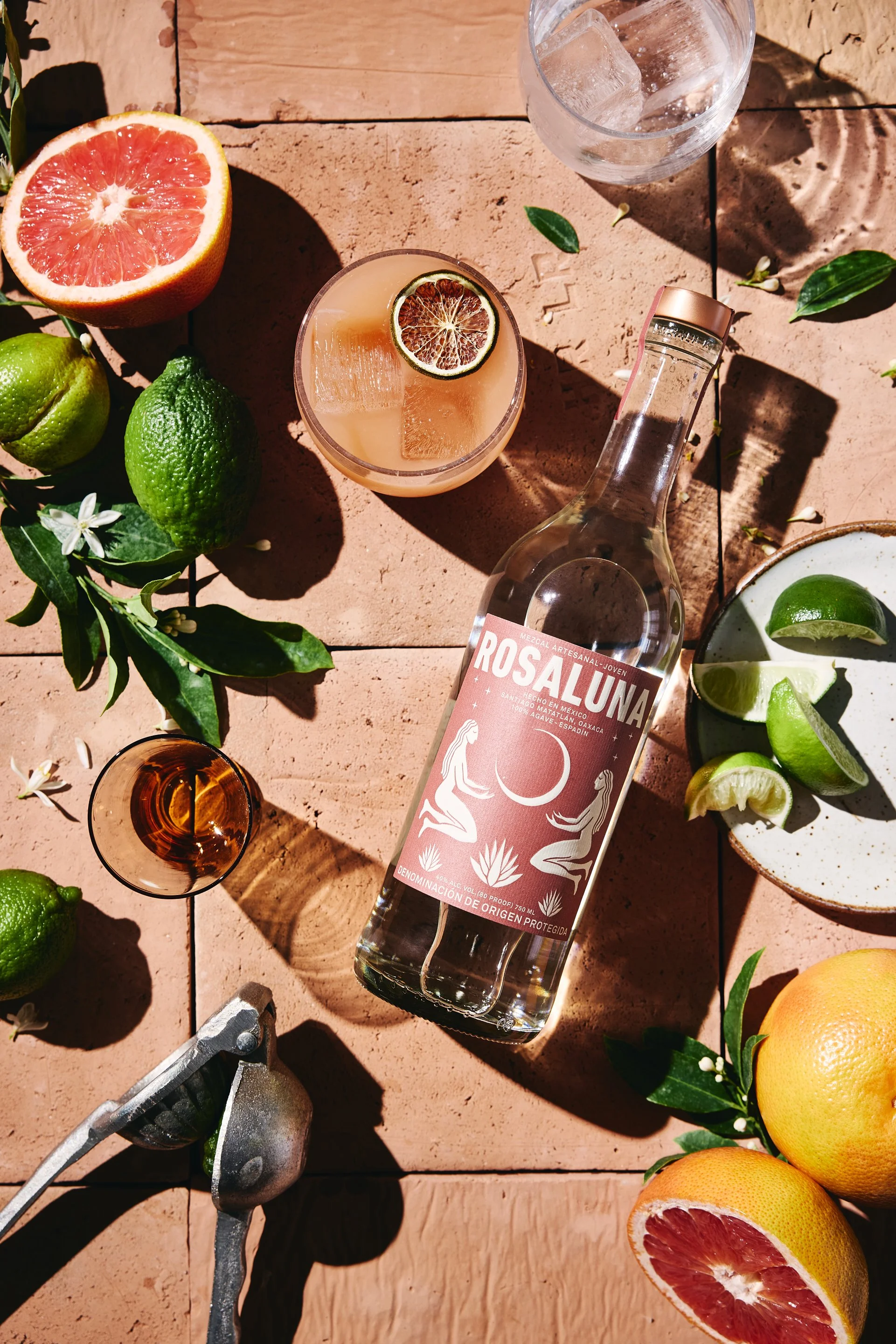 A bottle of Rosaluna mezcal surrounded by citrus fruits, a glass of mezcal with a grapefruit slice, a glass of water with ice, a plate with lime wedges, and citrus fruits on a tiled surface with sunlight and shadows.