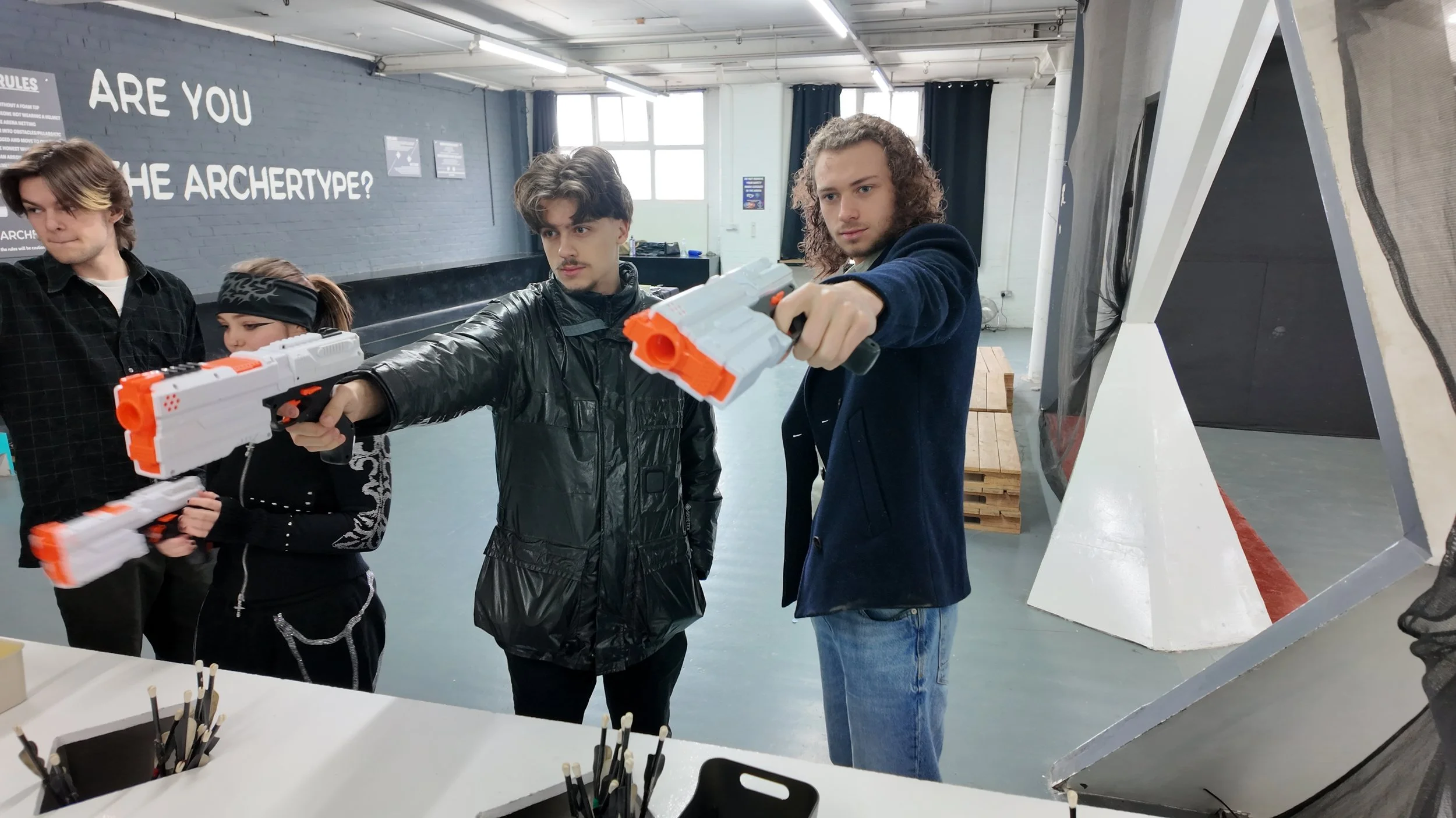 Four teenagers, three males and one female, are inside an indoor laser tag arena. They are holding laser guns and appear to be in a game. The arena has modern decor, with a large gray structure on the right and a wall with text and windows in the background.